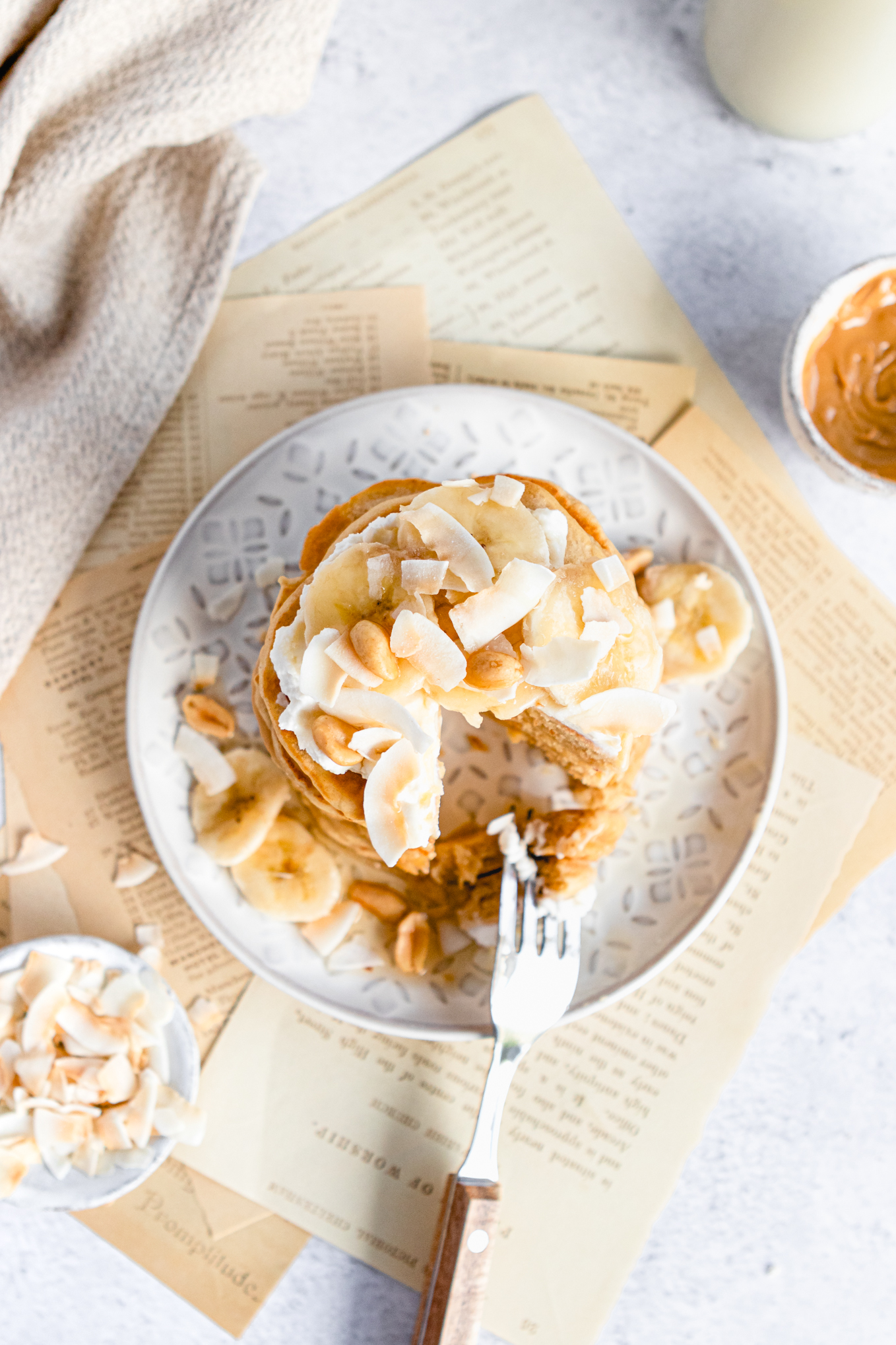 Peanut butter pancakes cut into with a fork on a plate. Side bowl of peanut butter and coconut flakes. 